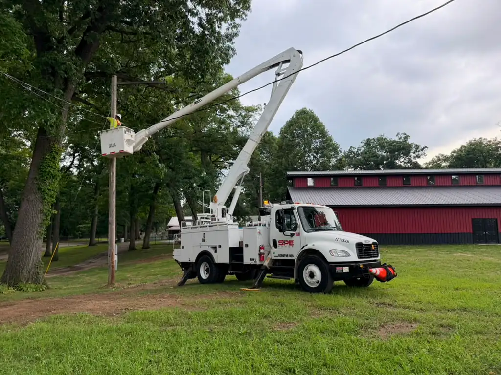 SPE bucket truck on grass with a lineman working at a wood pole, a red barn behind